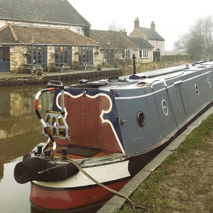 A narrowboat is moored at a canal bank, opposite an English country pub